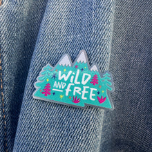 Load image into Gallery viewer, An enamel pin on a jean jacket. The pin features the words &quot;wild and free&quot; inside illustrated mountains with illustrated trees. 