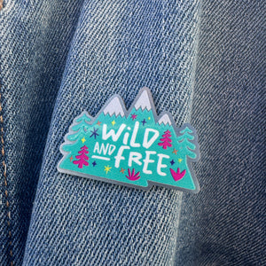 An enamel pin on a jean jacket. The pin features the words "wild and free" inside illustrated mountains with illustrated trees. 