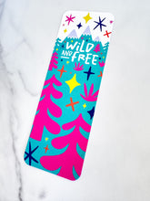 Load image into Gallery viewer, A bookmark placed on a tabletop. The bookmark features the words &quot;wild and free&quot; inside illustrated snow-capped mountains with illustrated trees. 