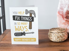 Load image into Gallery viewer, A card on a wood tabletop and on the right side of the card is a woven basket, a pink plant pot with a cactus in it and a pink crystal rock. The card features the words “You Help Fix Things in so Many Ways, Happy Father&#39;s Day” with an illustrated hammer and screwdriver around the words. 