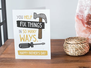 A card on a wood tabletop and on the right side of the card is a woven basket, a pink plant pot with a cactus in it and a pink crystal rock. The card features the words “You Help Fix Things in so Many Ways, Happy Father's Day” with an illustrated hammer and screwdriver around the words. 