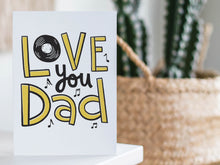 Load image into Gallery viewer, A card on a wood tabletop with an object in the background that is out of focus. The card features the words “Love you Dad” with a vinyl record as the “O” of love and music notes around the letters. 