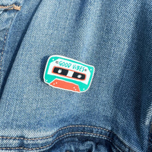 Load image into Gallery viewer, An enamel pin featuring the words &quot;good vibes&quot; inside an illustrated cassette tape. The pin is on a jean jacket.