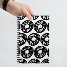 Load image into Gallery viewer, A spiral notebook being held by a person. The white spiral notebook has black spiral binding. The pattern design features the words &quot;good vibes&quot; inside an illustrated vinyl record.  