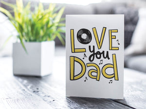 A greeting card is featured on a wood coffee table with a green plant in a white planter in the background. The card features the words  “Love you Dad” with a vinyl record as the “O” of love and music notes around the letters. 