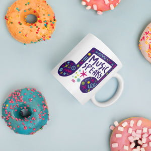 A white mug laying on a tabletop with doughnuts around it. The mug features a design with the words 'music speaks' in an illustrated musical note with sketchy elements around it.