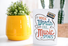 Load image into Gallery viewer, A greeting card is on a table top with a yellow plant pot and a green plant inside. The card features the words “When words fail, music speaks.”