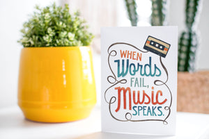 A greeting card is on a table top with a yellow plant pot and a green plant inside. The card features the words “When words fail, music speaks.”