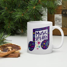 Load image into Gallery viewer, A white mug on a tabletop with part of a pine tree in the background. The mug features a design with the words &#39;music speaks&#39; in an illustrated musical note with sketchy elements around it.