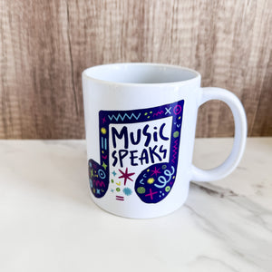 A white ceramic mug on a tabletop. The mug features a design with the words 'music speaks' in an illustrated musical note with sketchy elements around it.