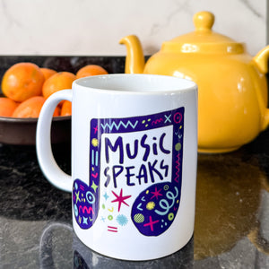 A white mug on a black countertop with a tea pot and oranges behind it. The mug features a design with the words 'music speaks' in an illustrated musical note with sketchy elements around it.