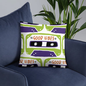 A white pillow on a blue sofa. The white pillow features the artwork on a white background with an illustrated cassette tape pattern with the words 'good vibes.'