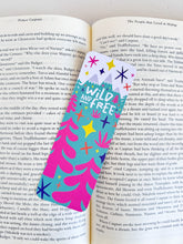 Load image into Gallery viewer, A bookmark placed inside a book. The bookmark features the words &quot;wild and free&quot; inside illustrated snow-capped mountains with illustrated trees. 