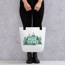 Load image into Gallery viewer, A person holding a white tote bag with black handles. The tote bag has the design with the words &#39;wild and free&#39; with the words inside illustrated mountains with illustrated trees around the words. 