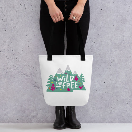 A person holding a white tote bag with black handles. The tote bag has the design with the words 'wild and free' with the words inside illustrated mountains with illustrated trees around the words. 