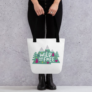 A person holding a white tote bag with black handles. The tote bag has the design with the words 'wild and free' with the words inside illustrated mountains with illustrated trees around the words. 