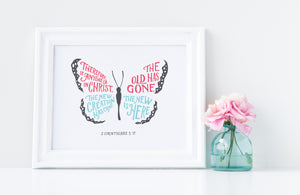 Artwork in a white frame with the with a white matte. The frame is leaning on a white counter with a pink flower in a blue case next to it. The hand drawn artwork features the scripture verse "Therefore, if anyone is in Christ, the new creation has come. The old has gone. The new is here." The words are featured inside a butterfly. 