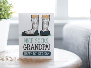 A card on a wood tabletop with an object in the background that is out of focus. The card features the words “Nice Socks Grandpa, Happy Father’s Day” with an illustrated of legs with patterned socks and shoes. 