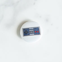 Load image into Gallery viewer, A button pin on a white tabletop. The button features an illustration of a retro Nintendo-like game controller. 