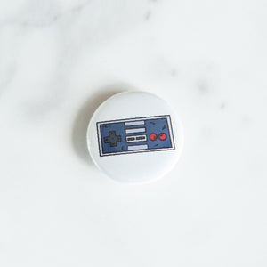 A button pin on a white tabletop. The button features an illustration of a retro Nintendo-like game controller. 