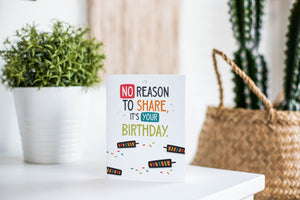 A greeting card is featured on a white tabletop with a white planter in the background with a green plant. There’s a woven basket in the background with a cactus inside. The card features the words “No reason to share it’s your birthday!”