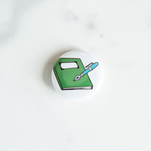 Load image into Gallery viewer, A buttons on a white tabletop. The button features an illustration of a notebook and pen.