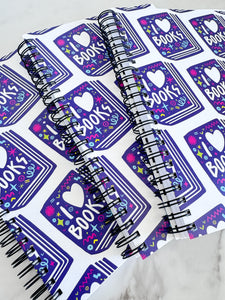 A closeup of three spiral notebooks are on a tabletop. The white spiral notebooks have black spiral binding. The pattern design features the words "I love books" inside an illustrated book with the word love as a heart. 