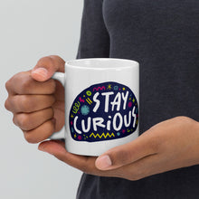 Load image into Gallery viewer, A person holding a white mug. The mug features a design with the words &#39;stay curious&#39; in an illustrated oval shape with sketchy elements around it. 