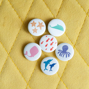 A set of six buttons on a yellow background. The buttons feature illustrations of whales, fish, starish, dolphins, and an octopus. 