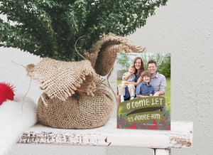 A photo of a one-sided Christmas card showing the front of the card standing up next to the bottom of a small Christmas tree with the base wrapped in burlap. The photo card features one photo with the words “O Come Let Us Adore Him” below with illustrated red leaves.