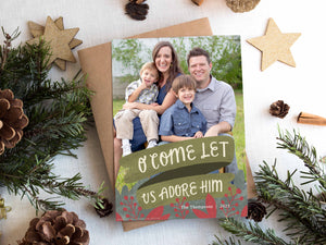 A photo of a one-sided Christmas card showing the front of the card on top of a brown wrapped gift on a white tabletop. Around the gift are pine needles, pinecones and wood star ornaments. The photo card features one photo with the words “O Come Let Us Adore Him” below with illustrated red leaves.