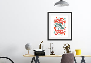 A black frame above a desk with artwork printed on white paper. The artwork features hand drawn lettering with the phrase "Great things never come from comfort zones."