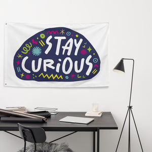 A large flag featured on a wall above a desk. The white flag features the words "stay curious" featured inside a a purple shape with sketchy illustrations around. 