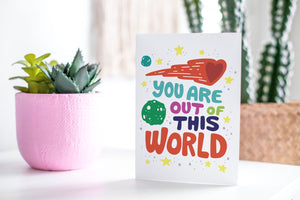 A greeting card featured standing up on a white tabletop with a pink plant pot in the background and some succulents in the pot. There’s a woven basket in the background with a cactus inside. The card features the words "You are out of this world” with space themed illustrations.