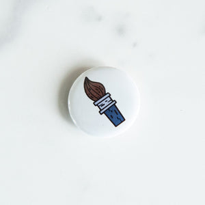 A button pin on a white tabletop. The button features an illustration of a paintbrush. 