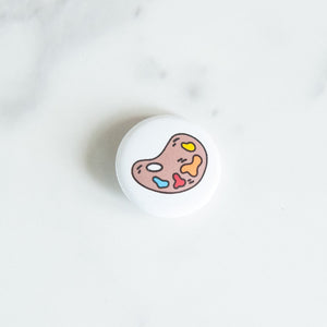 A button pin on a white tabletop. The button features an illustration of a paint palette. 