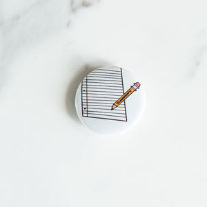 A buttons on a white tabletop. The button features an illustration of a pencil and paper.