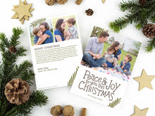 Load image into Gallery viewer, A photo of a two-sided Christmas card showing the front of the card on top of a brown wrapped gift on a white tabletop. Around the gift are pine needles, pinecones and wood star ornaments. The front of the card features a photo on the top portion and the words “Peace &amp; Joy to You This Christmas” with space to put your family name below. The back side of the card features two photos with space to write a yearly update. 