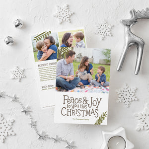 A photo of a double-sided Christmas card showing the front and back of the card laying on a white surface. Around the two sides of the card are surrounded with Christmas items. The front of the card features a photo on the top portion and the words “Peace & Joy to You This Christmas” with space to put your family name below. The back side of the card features two photos with space to write a yearly update. 