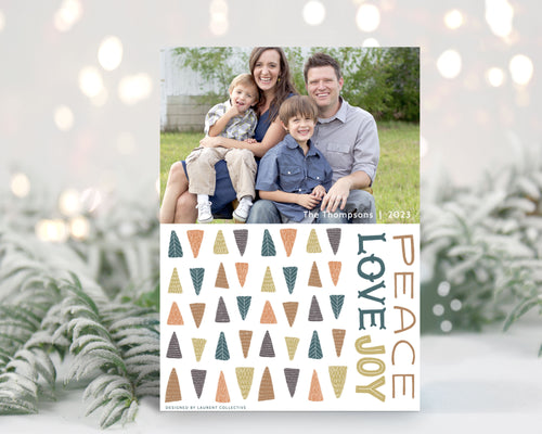 A photo of a one-sided Christmas card showing the front of the card standing up with pine needles behind and blurred white Christmas lights. The photo card features one photo with illustrated modern Christmas trees below. The words “Peace Love Joy” are to the left of the tree pattern. 