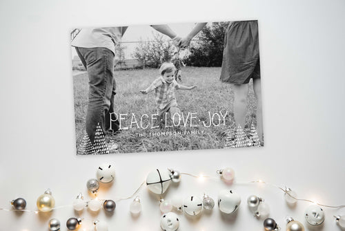 A photo of a one-sided Christmas card showing the front and back of the card laying on a white surface. To the bottom of the cards are silver and white small ornaments. The card features a photo with the words “Peace Love Joy” over the photo and a space to put your family name below. There are illustrated modern trees on the bottom. 