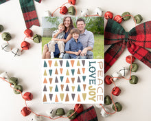 Load image into Gallery viewer, A photo of a one-sided Christmas card showing the front of the card laying on white table top. The card is surrounded by plaid red and green ribbon and red, green and white bells. The photo card features one photo with illustrated modern Christmas trees below. The words “Peace Love Joy” are to the left of the tree pattern. 
