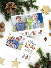 Load image into Gallery viewer, A photo of a two-sided Christmas card showing the front of the card on top of a brown wrapped gift on a white tabletop. Around the gift are pine needles, pinecones and wood star ornaments. The front of the card features a photo on the left side with the words “Peace Love Joy” with modern illustrated leaves below. The back of the card features two photos with the modern illustrated leaves. 