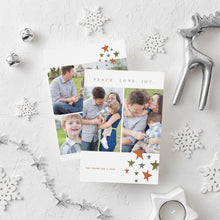 Load image into Gallery viewer, A photo of a double-sided Christmas card showing the front and back of the card laying on a white surface. Around the two sides of the card are surrounded with Christmas items. The front of the card features two photos with the words “Peace. Love. Joy.” above the photos. Below the photos are colored stars and a place where you can put your family name. The back of the card features three photos and colored stars. 