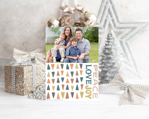 A photo of a one-sided Christmas card showing the front of the card standing up with Christmas items behind the card. There’s a sparkly star, glitter wrapped gift, ribbon and ornaments with a small silver tree around the photo card. The photo card features one photo with illustrated modern Christmas trees below. The words “Peace Love Joy” are to the left of the tree pattern. 