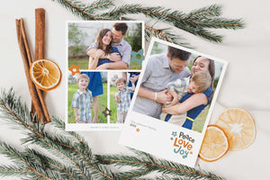 A photo of a double-sided Christmas card showing the front and back of the card laying on a white surface. Around the two sides of the card are pine needles, cinnamon sticks and dried oranges. The front of the card features a photo with the words “Peace Love joy” to the bottom right and space to the bottom left to put your family names. The words feature some modern illustrated flowers around it. The back of the card features three photos with modern illustrated flowers.