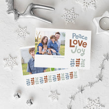 Load image into Gallery viewer, A photo of a double-sided Christmas card showing the front and back of the card laying on a white surface. Around the two sides of the card are surrounded with Christmas items. The front of the card features a photo on the left side with the words “Peace Love Joy” with modern illustrated leaves below. The back of the card features two photos with the modern illustrated leaves. 