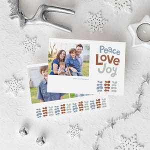 A photo of a double-sided Christmas card showing the front and back of the card laying on a white surface. Around the two sides of the card are surrounded with Christmas items. The front of the card features a photo on the left side with the words “Peace Love Joy” with modern illustrated leaves below. The back of the card features two photos with the modern illustrated leaves. 