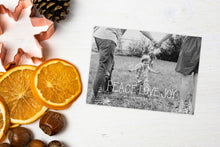 Load image into Gallery viewer, A photo of a Christmas card showing the front and back of the card laying on a white surface. Left of the card is a cookie cutter, pinecone, nuts and dried oranges. The card features a photo with the words “Peace Love Joy” over the photo and a space to put your family name below. There are illustrated modern trees on the bottom. 