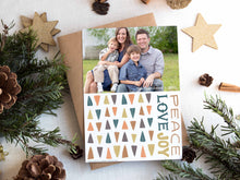 Load image into Gallery viewer, A photo of a one-sided Christmas card showing the front of the card on top of a brown wrapped gift on a white tabletop. Around the gift are pine needles, pinecones and wood star ornaments. The photo card features one photo with illustrated modern Christmas trees below. The words “Peace Love Joy” are to the left of the tree pattern. 